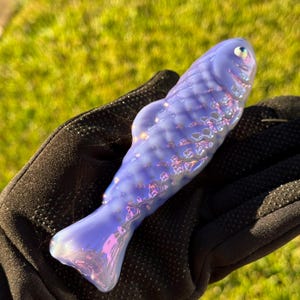 May include: A lavender-colored glass fish sculpture with iridescent highlights. The fish has textured scales and a rounded body. The sculpture is held in a black glove against a backdrop of green grass.
