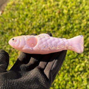 May include: A pink glass fish-shaped object with iridescent highlights. The fish has a round opening and a detailed scale pattern. The object is held in a black gloved hand against a green background.