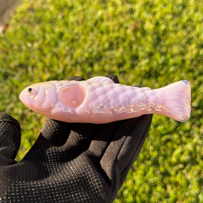 May include: A pink glass fish-shaped object with iridescent highlights. The fish has a round opening and a detailed scale pattern. The object is held in a black gloved hand against a green background.
