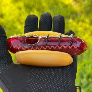 May include: A red glass pipe shaped like a hot dog with yellow and black accents. The pipe is sitting on a yellow glass bun.