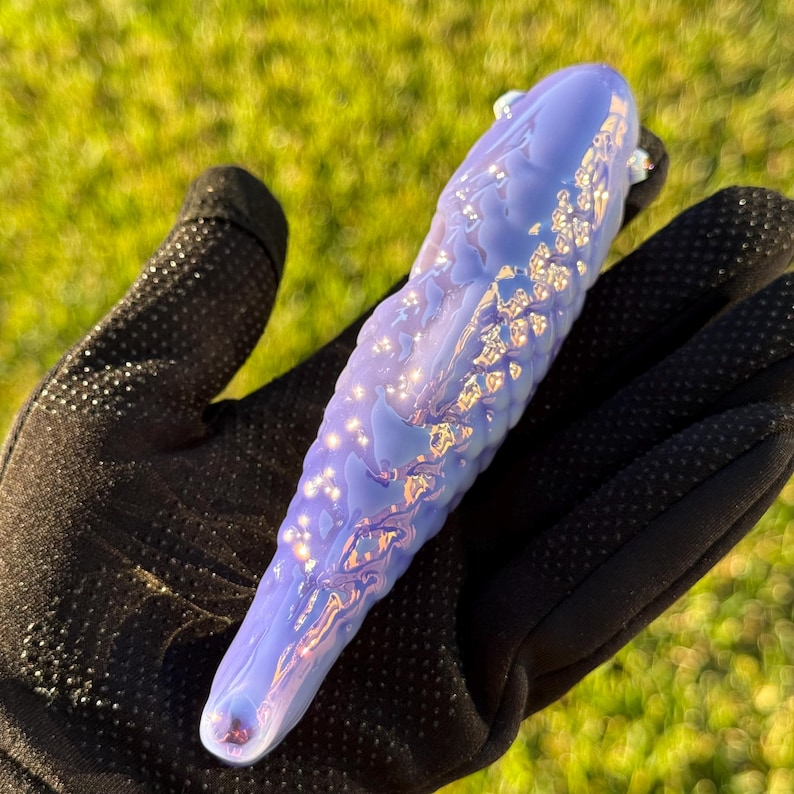 May include: A lavender-colored glass pipe with a textured surface and iridescent accents. The pipe is held in a black glove against a backdrop of green grass. The glass has a smooth, rounded shape with a flared end.