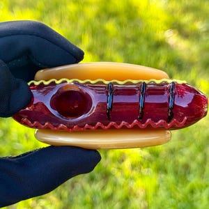 May include: A glass pipe shaped like a hot dog with a yellow and red design. The pipe is held in a hand wearing a black glove.