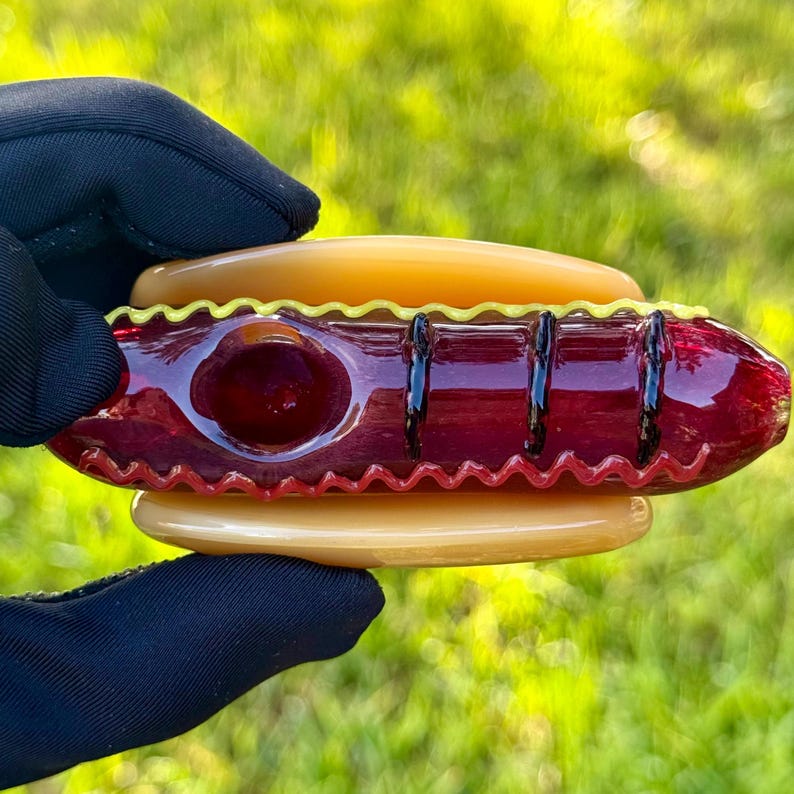 May include: A glass pipe shaped like a hot dog with a yellow and red design. The pipe is held in a hand wearing a black glove.