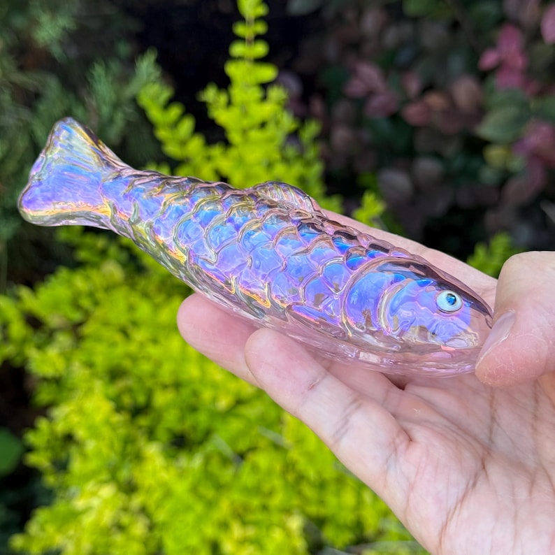 May include: A clear, iridescent glass fish figurine with a detailed scale pattern. The fish has a blue eye and a tail fin. The figurine is held in a hand against a blurred green and purple background.