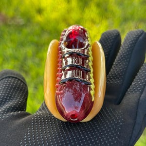 May include: A glass pipe shaped like a hot dog with a red and black design. The hot dog is in a yellow bun with a green and yellow mustard design.