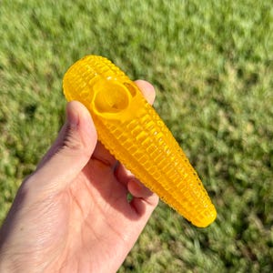 May include: A yellow glass pipe shaped like an ear of corn. The pipe has a bowl at the top and a long, narrow stem.