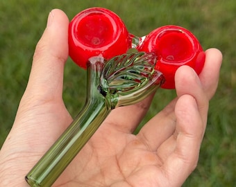 May include: A glass cherry pipe with a green stem and two red cherry bowls. The pipe is held in a hand.