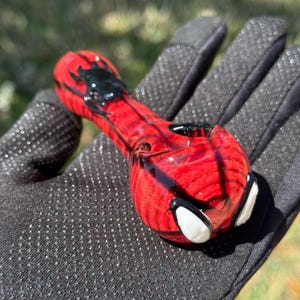 May include: A red and black glass pipe with a spider design. The pipe has a bowl, a stem, and a mouthpiece. The design includes a black spider on the top and white eye shapes on the end.