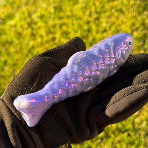 May include: A lavender-colored glass fish sculpture with iridescent highlights. The fish has a detailed scale pattern and a smooth, rounded body. The sculpture is held in a black glove against a blurred green background.