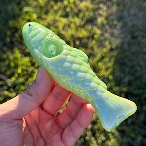 May include: A light green glass fish-shaped pipe. The pipe has a detailed scale pattern and a small opening on the top. The fish has a small, dark blue eye and a flared tail. The pipe is held in a hand.
