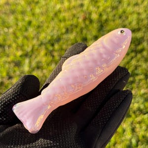 May include: A pink glass fish figurine with iridescent highlights. The fish has a detailed scale pattern and a small, round eye. The figurine is held in a black glove against a blurred green background.