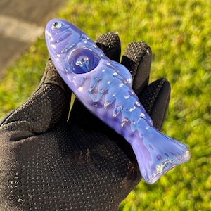 May include: A handcrafted, fish-shaped glass pipe in shades of blue and purple. The pipe features a detailed design with silver accents, a bowl, and a mouthpiece. The object is held in a black glove against a blurred green background.