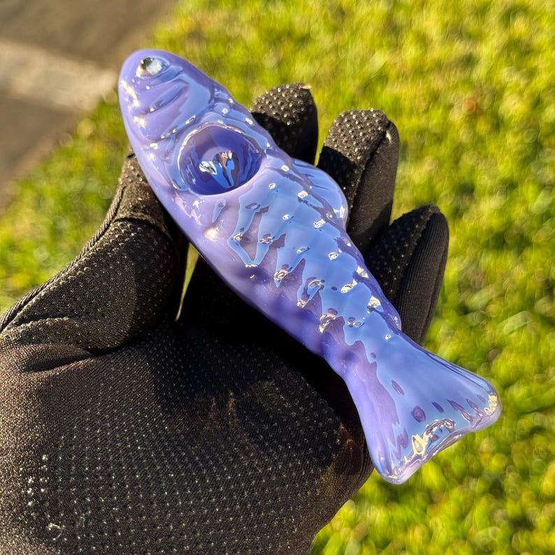 May include: A handcrafted, fish-shaped glass pipe in shades of blue and purple. The pipe features a detailed design with silver accents, a bowl, and a mouthpiece. The object is held in a black glove against a blurred green background.