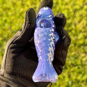 May include: A blue glass fish-shaped object with a textured surface. The object is held in a black glove. The fish has a tail and fins, and the body is a gradient of blue hues.