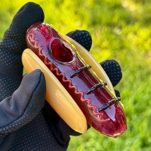 May include: A glass pipe shaped like a hot dog in a bun. The hot dog is red with black lines and a red and white zig-zag pattern. The bun is yellow with a white rim.