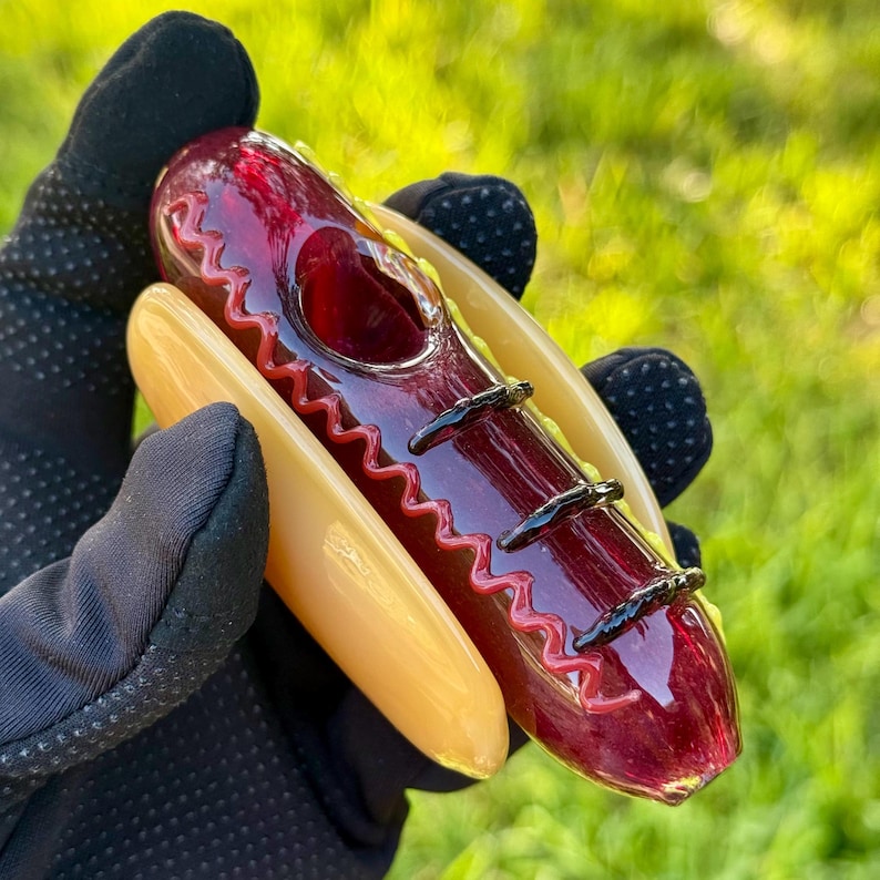 May include: A glass pipe shaped like a hot dog in a bun. The hot dog is red with black lines and a red and white zig-zag pattern. The bun is yellow with a white rim.
