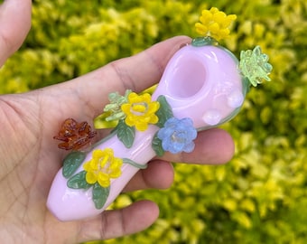 May include: A pink glass pipe adorned with colorful glass flowers and green leaves. The flowers are yellow, blue, and orange. The pipe is held in a hand against a blurred green background.