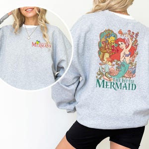 以下が含まれることがあります： スポーツグレーのスウェットシャツ。背中には、リトルマーメイドのアリエル、フランダー、セバスチャンのカラフルなグラフィックが描かれています。スウェットシャツの前面には、赤と黄色で「The Little Mermaid」の文字がプリントされています。クルーネックのスウェットシャツです。