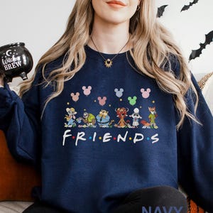 Pode incluir: Camisola azul marinho com um gr&aacute;fico colorido de personagens de desenhos animados e a palavra "FRIENDS" em branco. Acima dos personagens est&atilde;o formas de cabe&ccedil;a do Mickey Mouse em v&aacute;rias cores. A palavra "NAVY" &eacute; impressa na parte inferior.