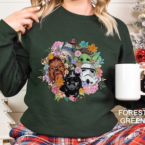 Puede incluir: Sudadera verde bosque con un dise&ntilde;o de corona floral con personajes de Star Wars, incluyendo a Chewbacca, Darth Vader, R2-D2, un Stormtrooper y Grogu. La sudadera tiene mangas largas y cuello redondo. El texto "FOREST GREEN" est&aacute; impreso en la parte inferior.