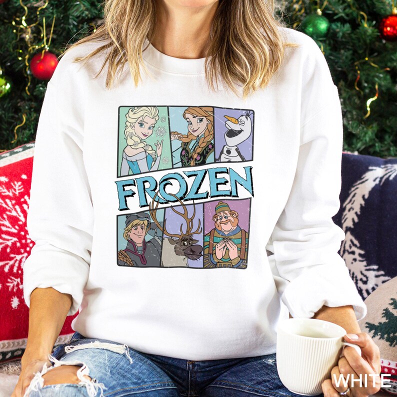 Puede incluir: Sudadera blanca con un gr&aacute;fico colorido de personajes de la pel&iacute;cula "Frozen". El gr&aacute;fico incluye a Elsa, Anna, Olaf, Kristoff, Sven y un hombre. La palabra "FROZEN" est&aacute; impresa en letras azules. La sudadera la lleva una persona.
