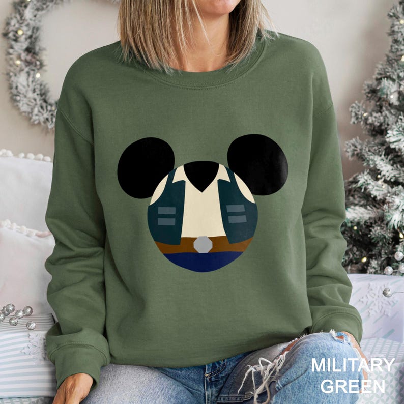 May include: Military green sweatshirt featuring a cartoon design. The design is a stylized character face with black ears, a white shirt, a blue jacket, and a brown belt. The text "MILITARY GREEN" is at the bottom.