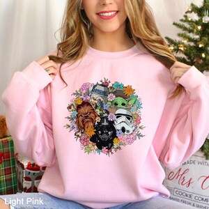 Puede incluir: Sudadera rosa claro con un dise&ntilde;o floral con personajes de Star Wars como Darth Vader, Chewbacca y Baby Yoda. El dise&ntilde;o est&aacute; rodeado de flores coloridas. El texto "Light Pink" es visible en la parte inferior.
