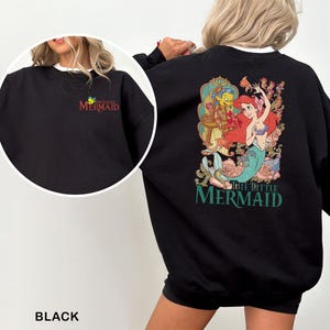 以下が含まれることがあります： 背中にリトルマーメイドのアリエルのカラフルなグラフィックが描かれた黒いスウェットシャツ。左胸には「The Little Mermaid」の文字が入った小さなロゴがあります。スウェットシャツは白い襟が付いています。