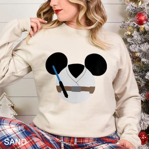 May include: Sand-colored sweatshirt featuring a cartoon design of a character's face with black ears, a white tunic, and a blue lightsaber. The design is a playful mashup of a famous character and a popular cartoon character.