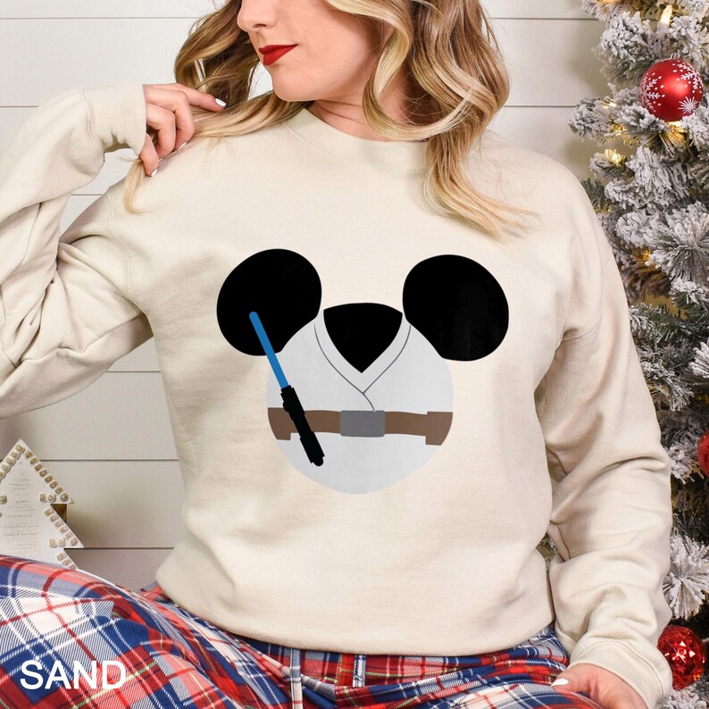 May include: Sand-colored sweatshirt featuring a cartoon design of a character's face with black ears, a white tunic, and a blue lightsaber. The design is a playful mashup of a famous character and a popular cartoon character.