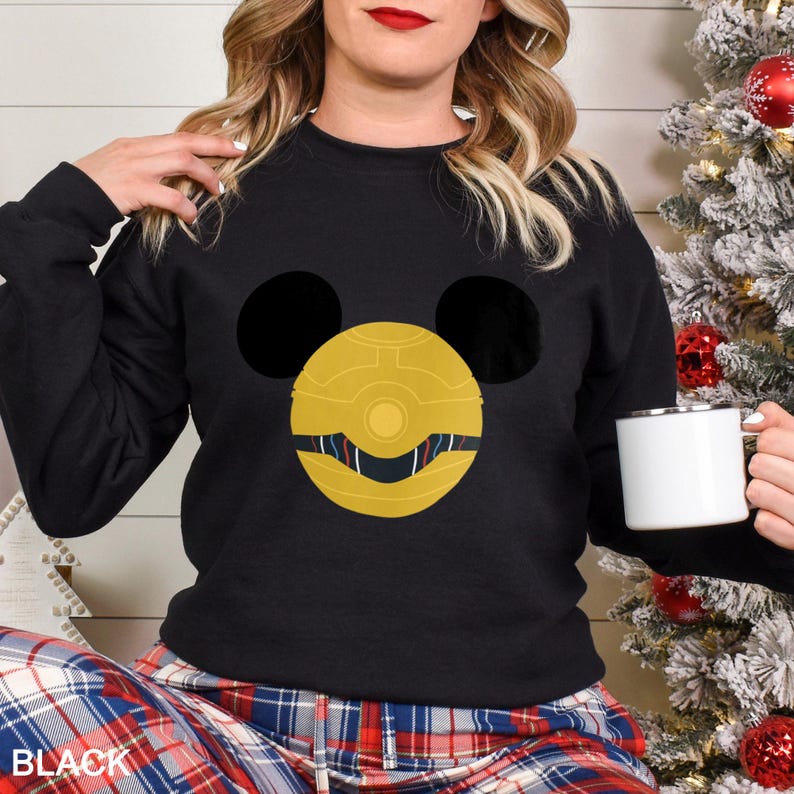 May include: Black sweatshirt featuring a gold and black graphic design. The design incorporates a circular element with a face-like appearance, flanked by two large black circles. The word "BLACK" is visible at the bottom left.