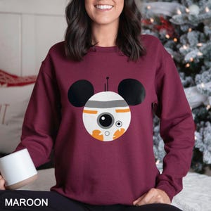 May include: Maroon sweatshirt featuring a BB-8 droid design with black Mickey Mouse ears. The droid has a white body with orange and gray details, and a large black eye. The word "MAROON" is printed at the bottom of the image.