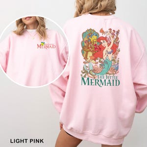以下が含まれることがあります： リトルマーメイドのアリエル、フランダー、セバスチャンが描かれたライトピンクのスウェットシャツ。「The Little Mermaid」の文字が前面にプリントされています。スウェットシャツの背面には、アリエル、フランダー、セバスチャンの大きなグラフィックがあります。