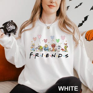 Pode incluir: Camisola branca com a palavra "FRIENDS" em preto. Acima da palavra, h&aacute; uma ilustra&ccedil;&atilde;o de desenho animado de personagens de animais segurando bal&otilde;es em forma de Mickey Mouse. A palavra "WHITE" &eacute; impressa em branco na parte inferior.
