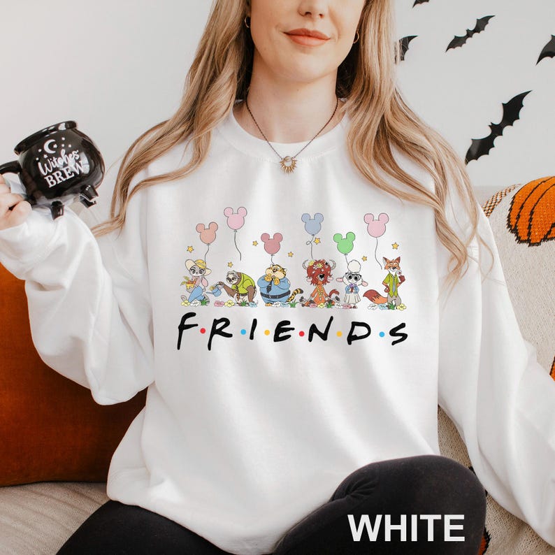 Pode incluir: Camisola branca com a palavra "FRIENDS" em preto. Acima da palavra, h&aacute; uma ilustra&ccedil;&atilde;o de desenho animado de personagens de animais segurando bal&otilde;es em forma de Mickey Mouse. A palavra "WHITE" &eacute; impressa em branco na parte inferior.