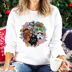 Puede incluir: Sudadera blanca con un dise&ntilde;o floral colorido que presenta personajes de Star Wars, incluyendo a Darth Vader, Chewbacca, R2-D2, un Stormtrooper y Grogu. El dise&ntilde;o est&aacute; rodeado de flores en tonos rosas, naranjas y azules.