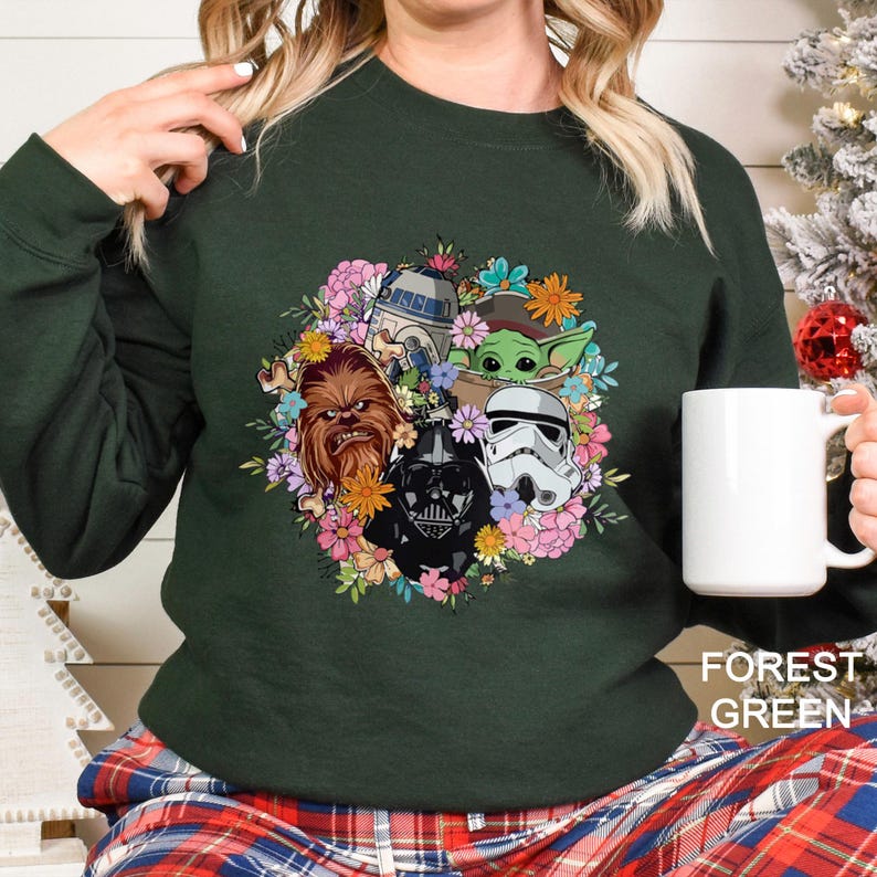 Puede incluir: Sudadera verde bosque con un dise&ntilde;o de corona floral con personajes de Star Wars, incluyendo a Chewbacca, Darth Vader, R2-D2, un Stormtrooper y Grogu. La sudadera tiene mangas largas y cuello redondo. El texto "FOREST GREEN" est&aacute; impreso en la parte inferior.