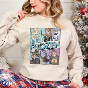 Puede incluir: Una sudadera de color arena con un gr&aacute;fico colorido de personajes de la pel&iacute;cula "Frozen". El gr&aacute;fico incluye a Elsa, Anna, Olaf, Kristoff, Sven y un personaje con un su&eacute;ter a rayas azules y verdes. La palabra "FROZEN" est&aacute; impresa en azul.