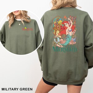 以下が含まれることがあります： ミリタリーグリーンのスウェットシャツ。リトルマーメイドのアリエル、フランダー、セバスチャンのグラフィックが特徴です。スウェットシャツの背面には、映画のタイトルが描かれたカラフルなイラストが描かれています。前面には、映画のタイトルが赤と黄色で表示されています。