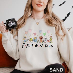 Pode incluir: Camisola cor de areia com um gr&aacute;fico de personagens de desenhos animados segurando bal&otilde;es em forma de Mickey Mouse. A palavra "FRIENDS" est&aacute; impressa em preto abaixo dos personagens. Um estilo casual e confort&aacute;vel.
