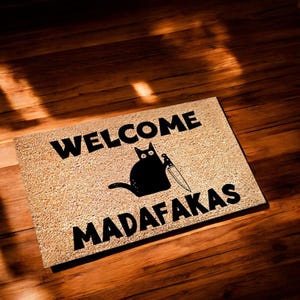 Könnte beinhalten: Eine braune Fußmatte mit dem Text "WELCOME MADAFAKAS" in schwarzen Buchstaben. Eine schwarze Katze mit einem Messer sitzt auf der Matte.