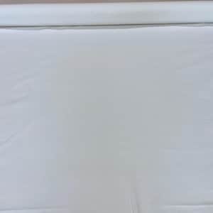 May include: A large roll of white fabric, unrolled and spread out on a surface. The fabric appears to be a solid, plain weave, with a smooth texture. The roll is visible in the background, indicating a continuous length of material.