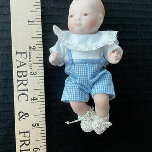 May include: A small porcelain doll wearing a blue and white checkered outfit with a white lace collar and crocheted booties. The doll is approximately 4 inches tall and is posed sitting with its legs crossed.