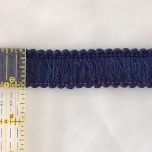 May include: Close-up of a navy blue fringe trim with a scalloped edge and a row of fringe. The trim is approximately 1 inch wide. A ruler is placed next to the trim for measurement.