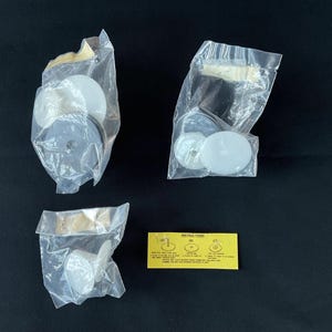 May include: An overhead shot shows three clear plastic bags containing various white and gray circular objects. A small yellow card with text and graphics is also present. The items are on a dark surface.