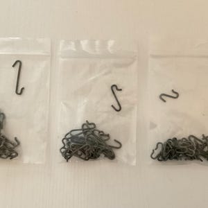 May include: Three clear plastic bags containing a large quantity of small, silver-colored metal hooks. Each bag contains a single larger hook.