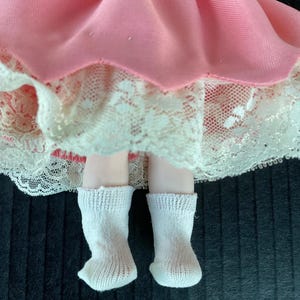 May include: A porcelain doll wearing a pink dress with white lace trim and white socks.