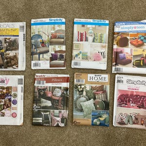 May include: A collection of seven sewing pattern envelopes, featuring designs for home decor items like pillows and cushions. The envelopes display various colors and images of finished products, with brand names like Simplicity and McCall's.