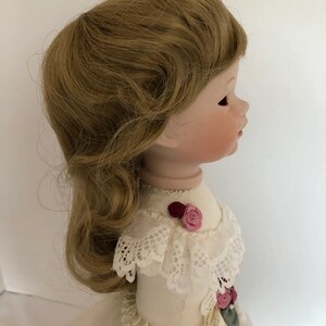 May include: A porcelain doll with long, wavy, light brown hair. The doll is wearing a white dress with lace trim and pink flower accents.