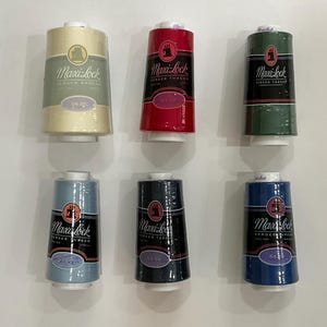 May include: Six spools of Maxi-Lock serger thread arranged in two rows. The top row features cream, red, and dark green spools. The bottom row includes light blue, dark gray, and royal blue spools. Each spool has a black label with white text.